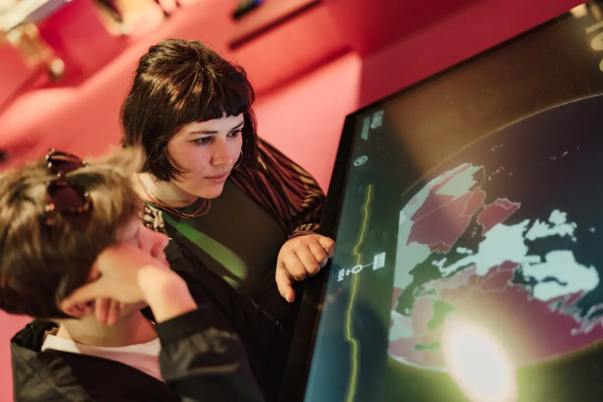 Zwei junge Frauen in einer Ausstellung schauen sich ein Exponat an. Das Exponat ist ein großer Bildschirm, auf dem eine geographische Karte zu sehen ist.