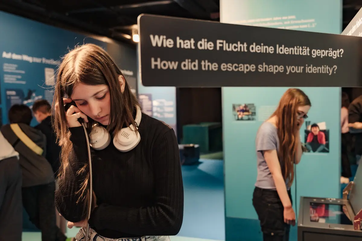 Eine Schülerin in einer Ausstellung. Die Person hört gerade über Kopfhörer etwas an. Auf einem Schild im Hintergrund steht "Wie hat die Flucht deine Identität geprägt?"
