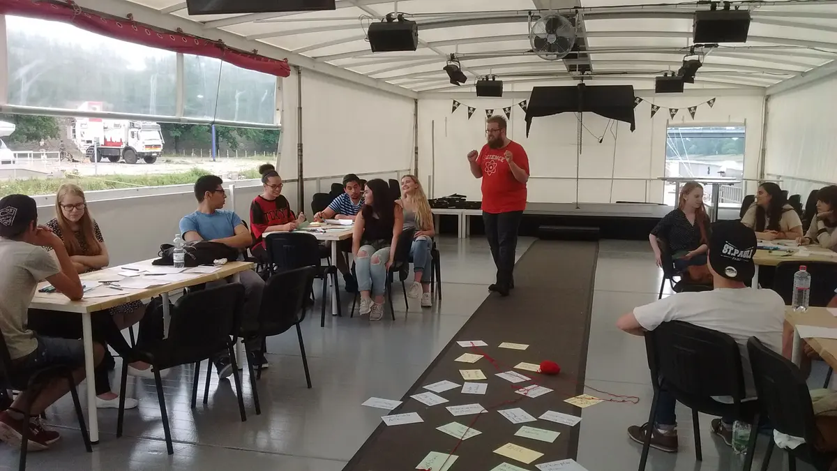 Jugendliche sitzen an mehreren Gruppentischen in einem Zelt. Auf dem Boden liegen viele Zettel verteilt. Eine Person im Hintergrund erklärt etwas.