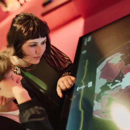 Zwei junge Frauen in einer Ausstellung schauen sich ein Exponat an. Das Exponat ist ein großer Bildschirm, auf dem eine geographische Karte zu sehen ist. 