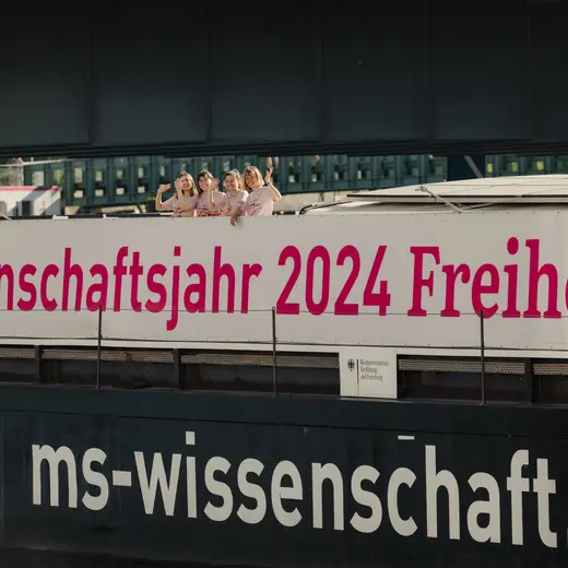 Fernaufnahme vom Schiff MS Wissenschaft. Auf der Schiffseite steht "Wissenschaftsjahr 2024 Freiheit". Am Deck winken aus der Ferne vier Lotsinnen der Kamera zu.