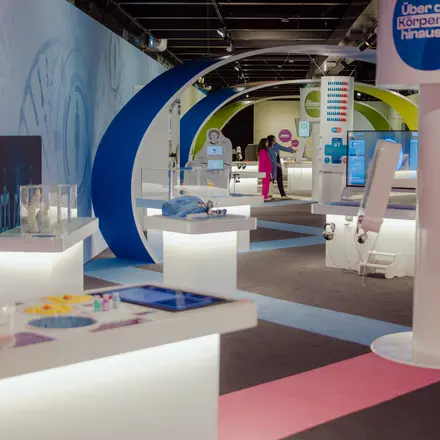Blick in den Ausstellungsraum der MS Wissenschaft. Man sieht weiße Tisch mit Exponaten sowie in der Mitte weiße Säulen, die Forschungsstationen. Der Raum wird gerahmt durch blaue Bögen aus Holz, die an eine DNA-Helix erinnern. 