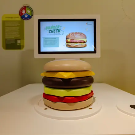 Ein Tisch, auf dem ein großer Burger aus Holz steht. Im Hintergrund ein Bildschirm an einer Wand, auf dem "Burger Check" steht. 