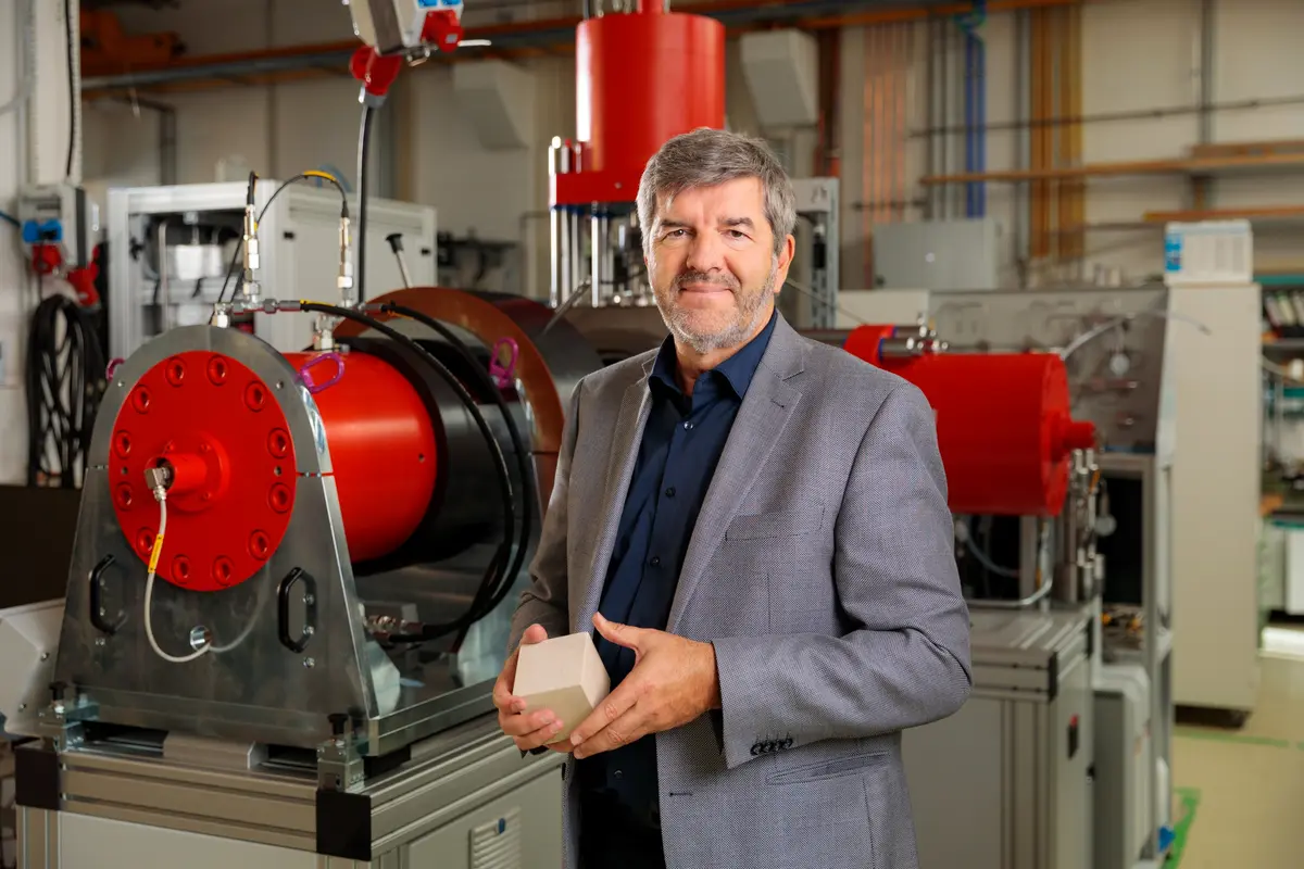 Eine Person mit einem blauen Tshirt und einem grauen Jacket steht vor einer Forschungseinrichtung, die aus roten und schwarzen dicken Rohren besteht.