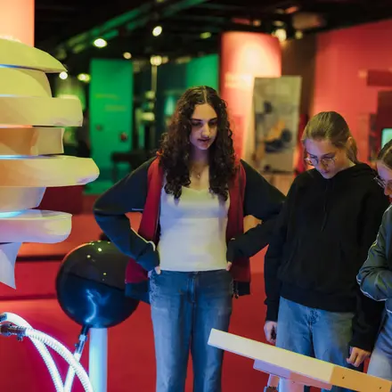 Ein Exponat- rechts eine große bewegliche Skulptur von einem weißen Kopf, der in Scheiben zerschnitten ist und ein Bildschirm davor. Vor dem Exponat stehen drei Besucherinnen. 