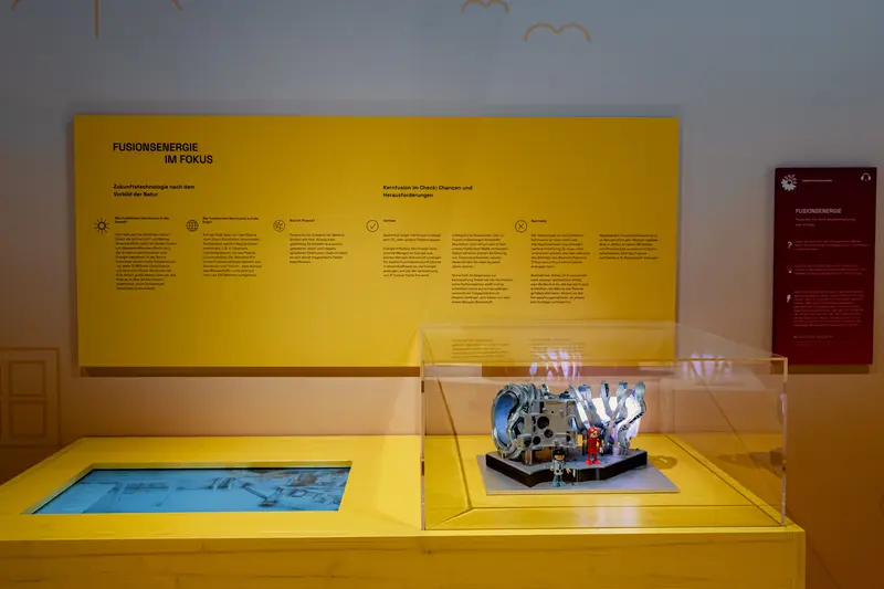 Das Bild zeigt den Exponatstisch mit einem Modell und einem Bildschirm und die gelbe Informationstafel im Hintergrund.