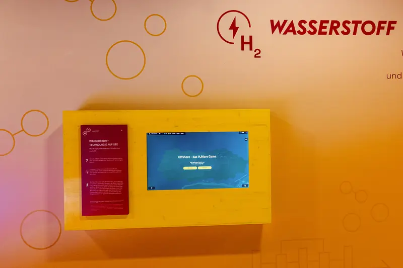 Das Bild zeigt einen an der Wand hängenden Bildschirm. Auf dem Screen kann das H2Mare Game gestartet werden.