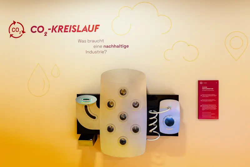 Das Bild zeigt das an der Wand hängende Exponat. Es ist grau und besteht aus drei Elementen.