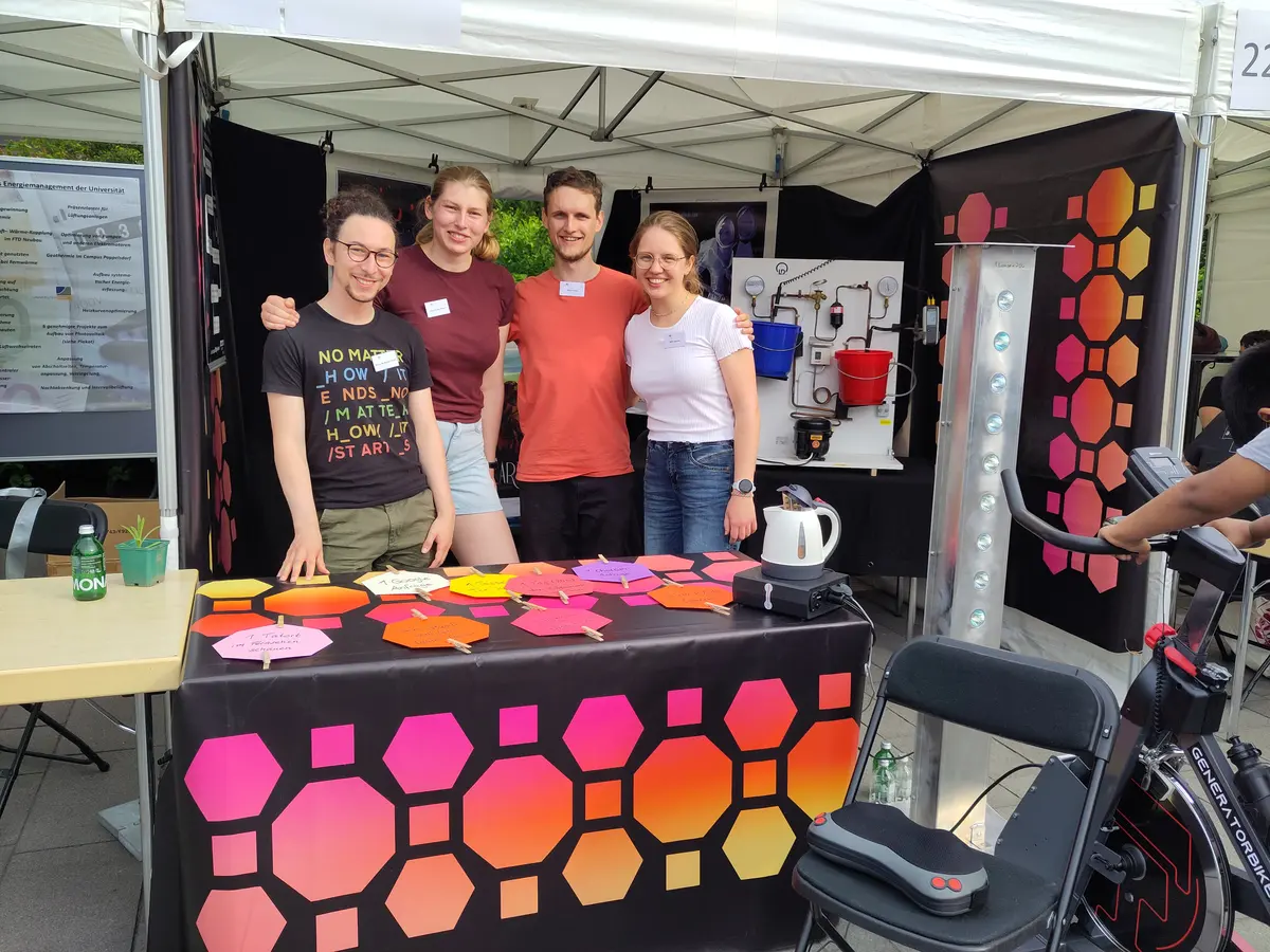 Drei Personen an einem Stand, rechts im Vordergrund ein Hometrainer.