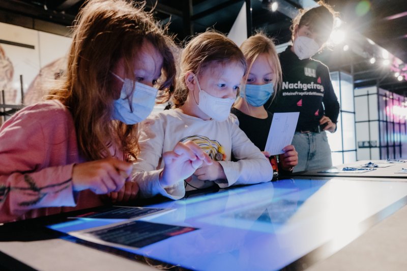 Drei Mädchen und eine Ausstellungslotsin betrachten einen Bildschirm, um Quallengalaxien ausfindig zu machen.