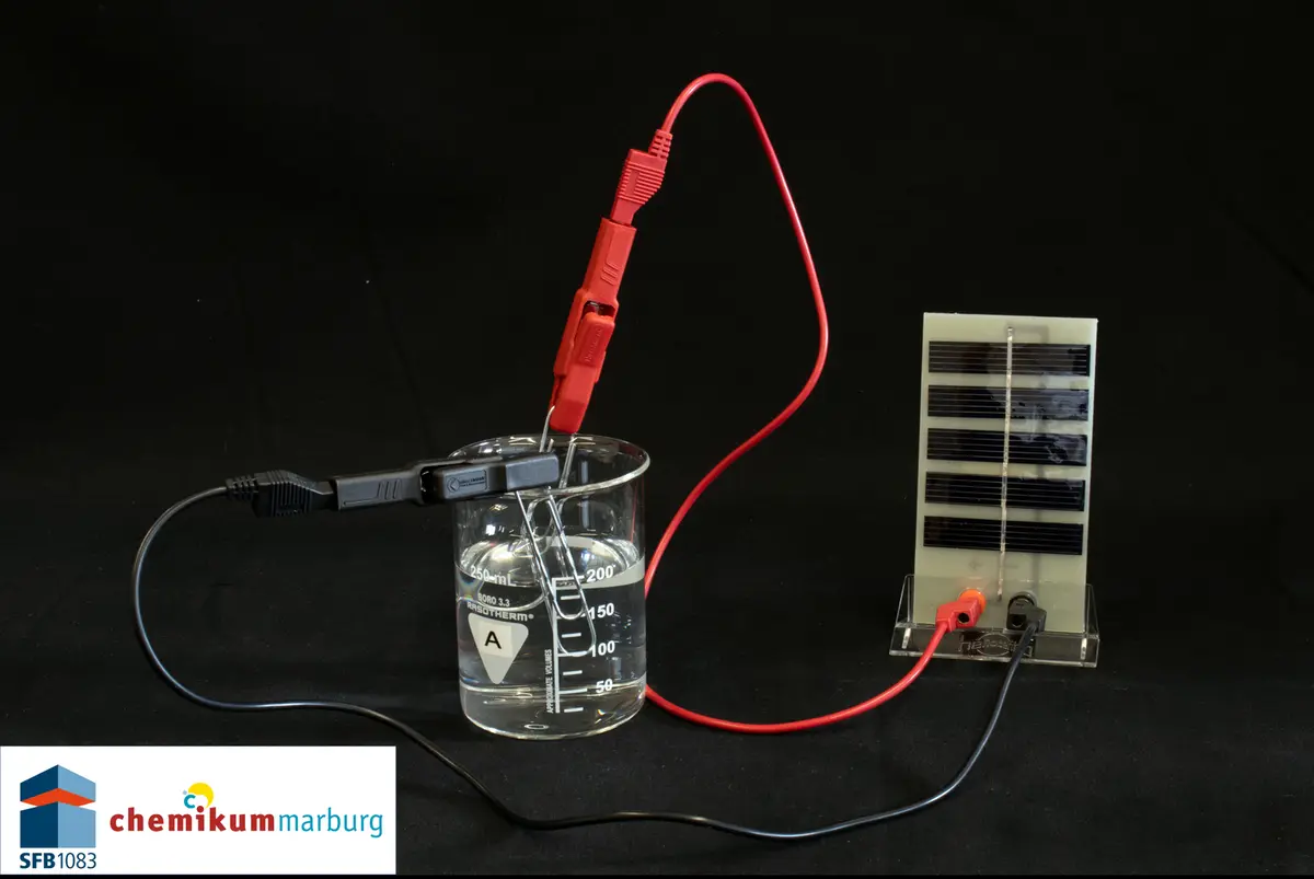 Ein Reagenzglas mit einer durchsichtigen Flüssigkeit und zwei Kabel drin, die an ein kleiner Solarpanel angeschlossen sind.